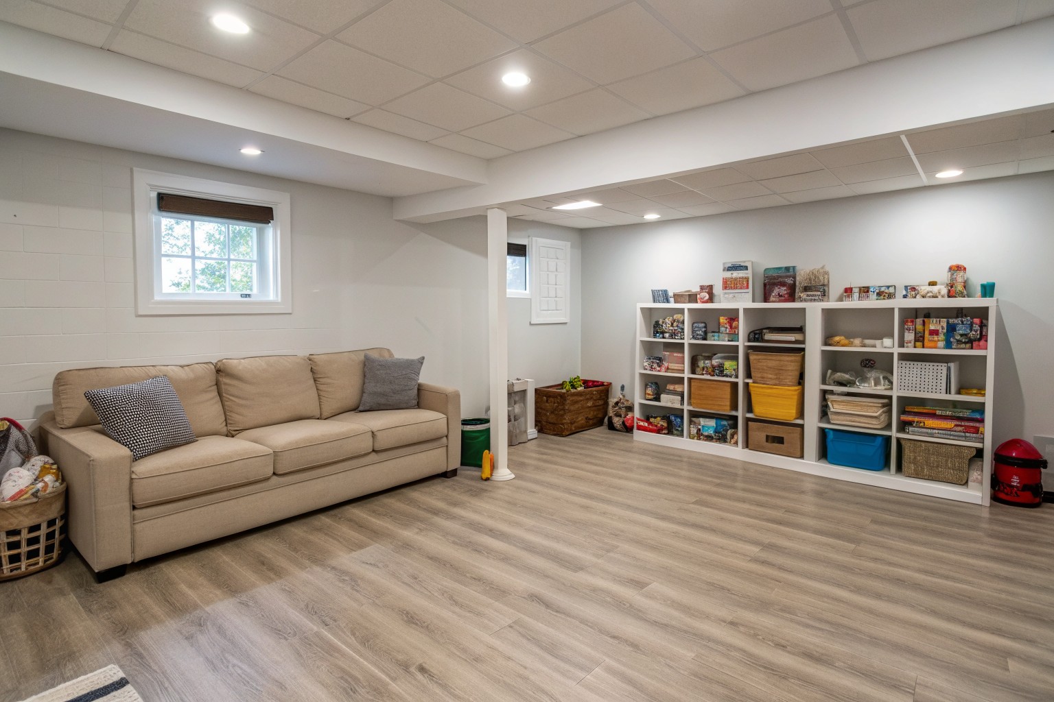 A dry finished basement playroom with a sectional sofa, toy shelves and a small egress window