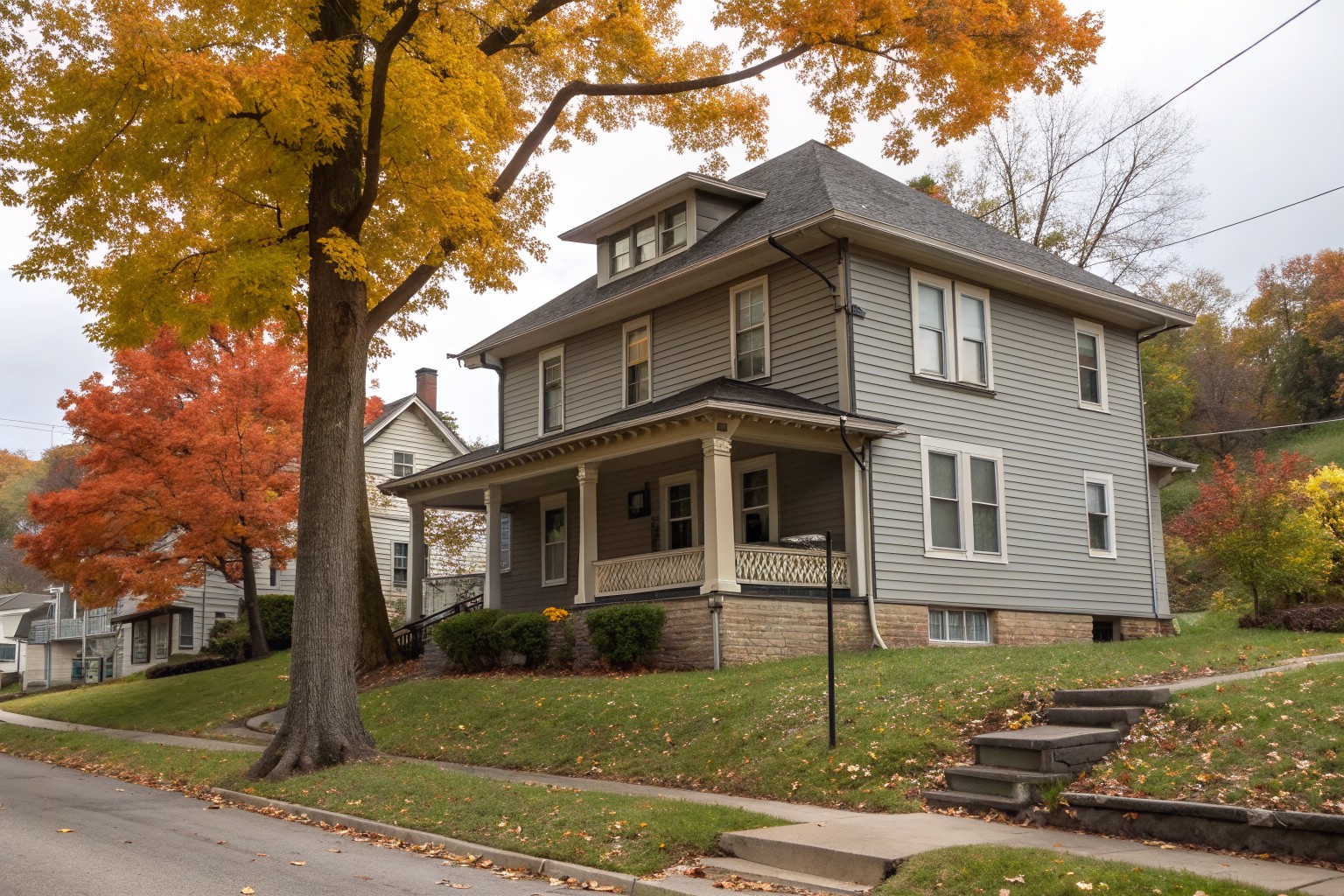 A 1920s gray clapboard foursquare with a wide porch and small basement windows in a sandstone block foundation on a hilly street